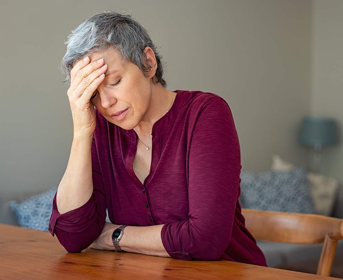 A woman sits at a dining room table, closing her eyes and resting her forehead in her hand.