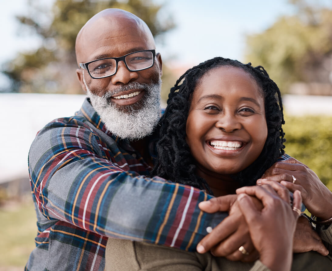 A man hugs a woman in a park. They both smile at the camera.