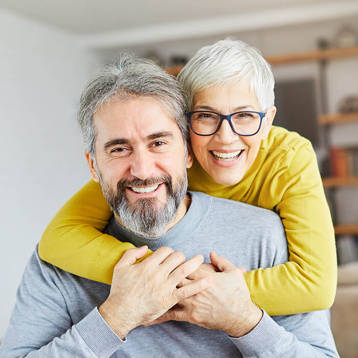 A woman stands behind a man, hugging him, while they both look at the camera.