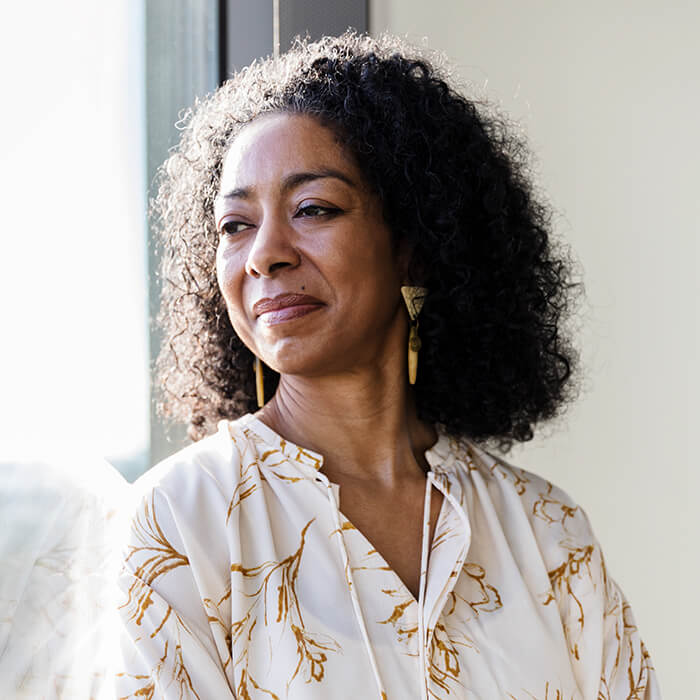 A woman stands in the light of a window, looking out and grinning.