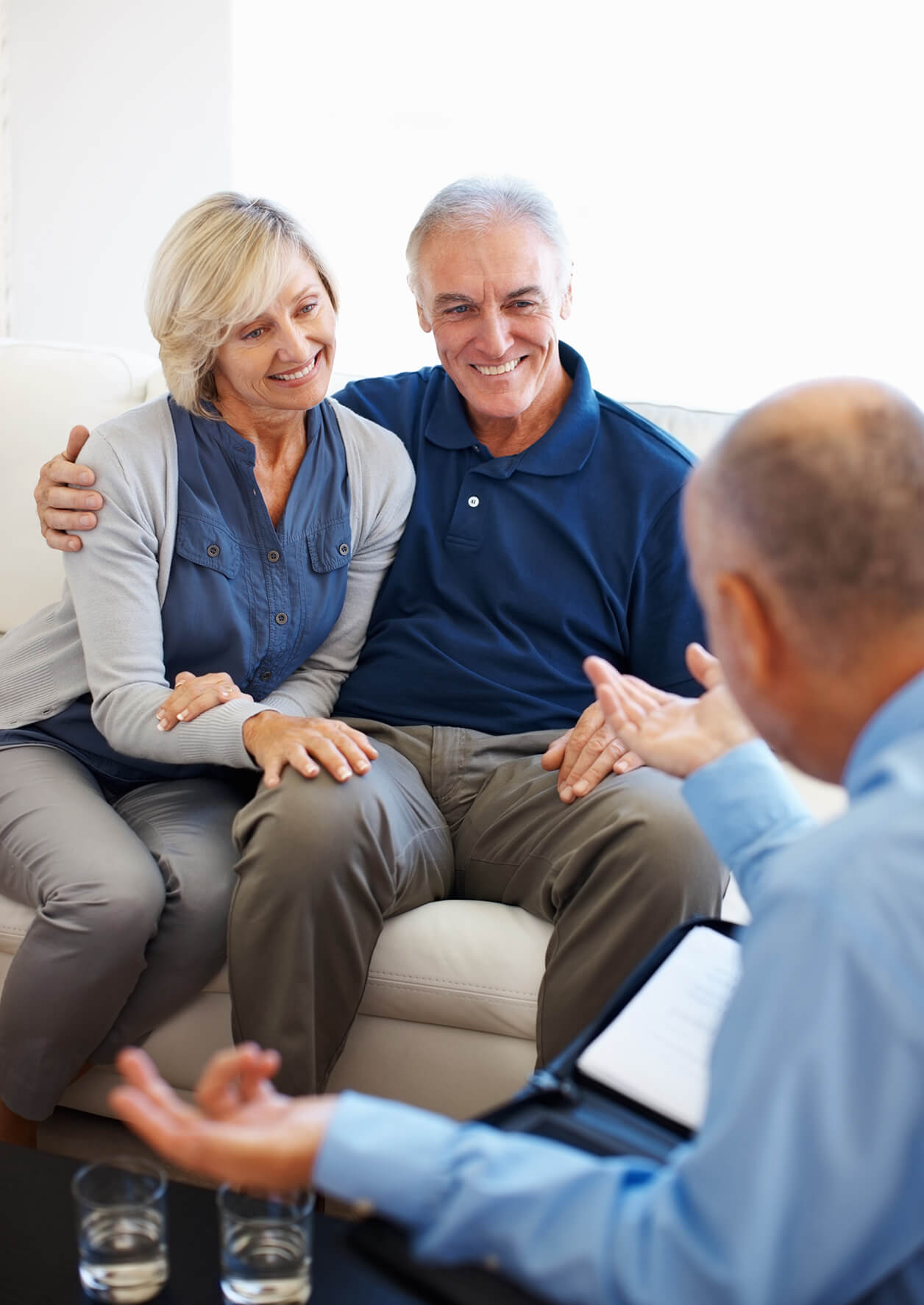 A couple sits on a couch talking to a man in a dress shirt.