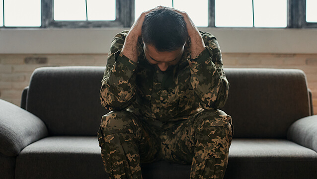 A man in military fatigues sits on a couch, looking down with his hands behind his neck. You can get Social Security Disability for PTSD.