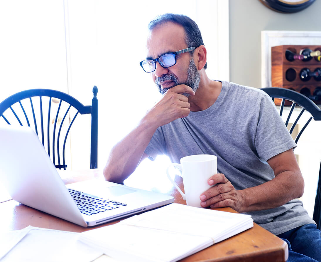 A man sits at a kitchen table with a coffee mug, looking at a laptop.