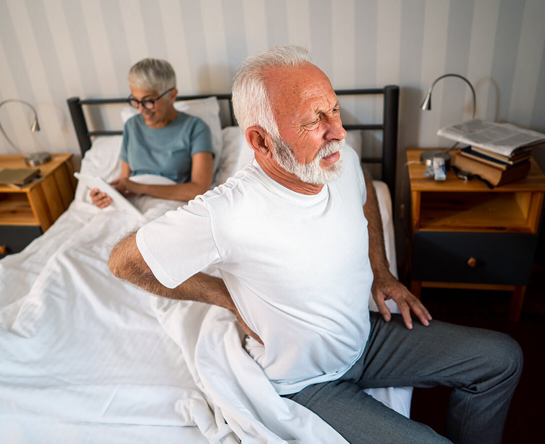 A man sits on the edge of a bed, holding his lower back, while a woman sits on the other side of the bed reading papers.