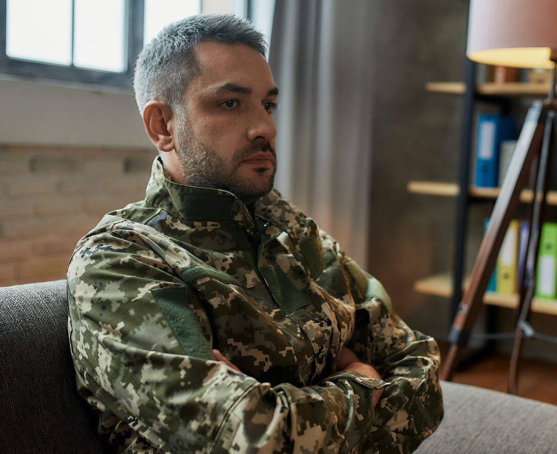 A man in military fatigues sits on a couch with his arms crossed.