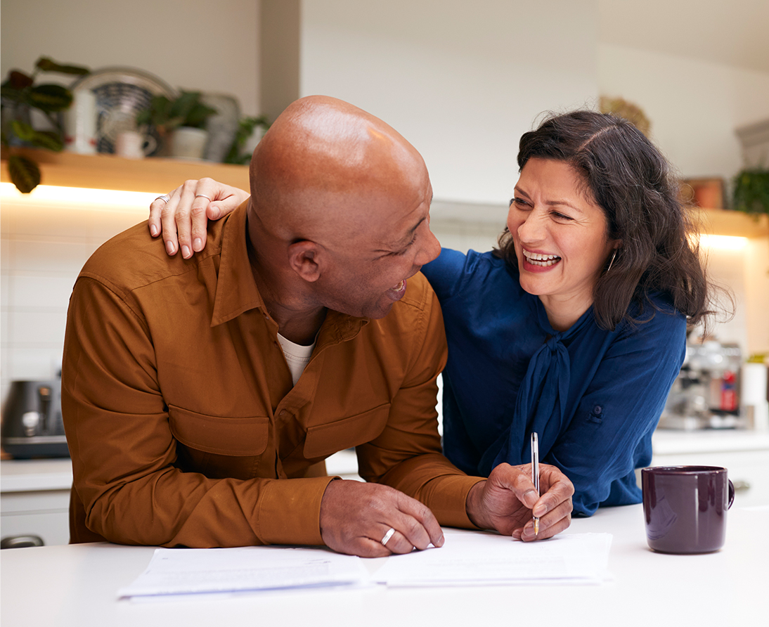 A woman puts her arm around a man's shoulder as they stand at a kitchen island over paperwork.