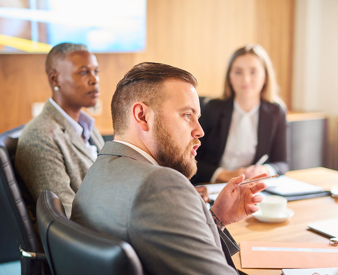 People sit around a conference room table, listening to a man speaking.