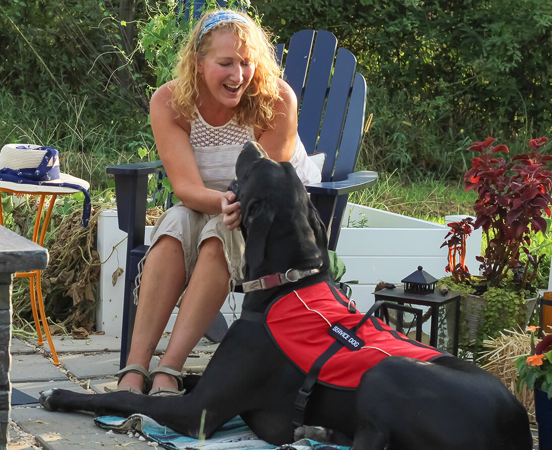 A woman sits on a patio chair, smiling and touching the face of a large, brown service dog.