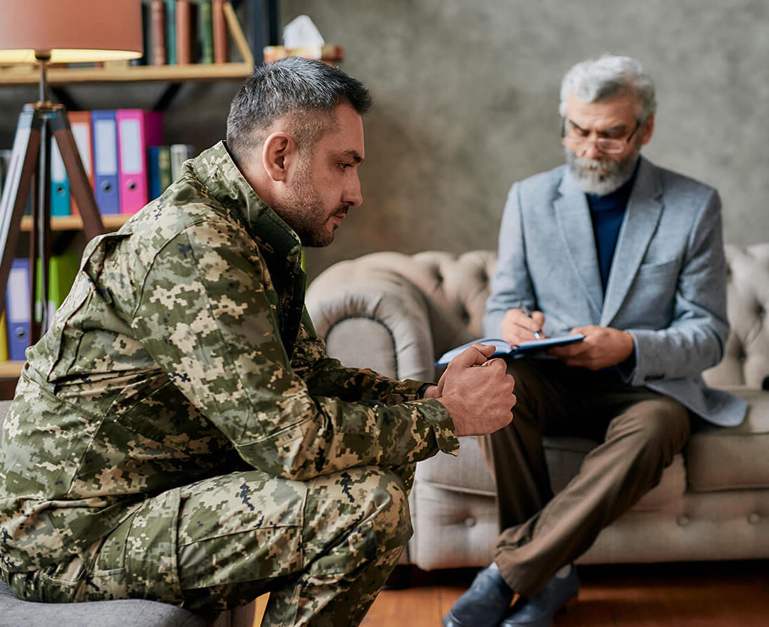 A man with a clipboard listens to a man in military fatigues sitting on a couch.