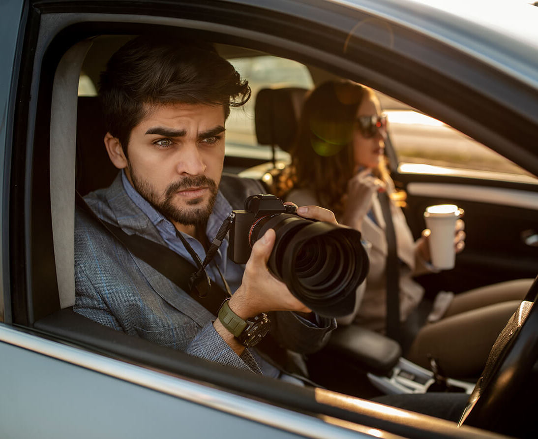 A man looks out the driver's side window of a car with a camera in hand, while a woman sits in the passenger seat holding a coffee.