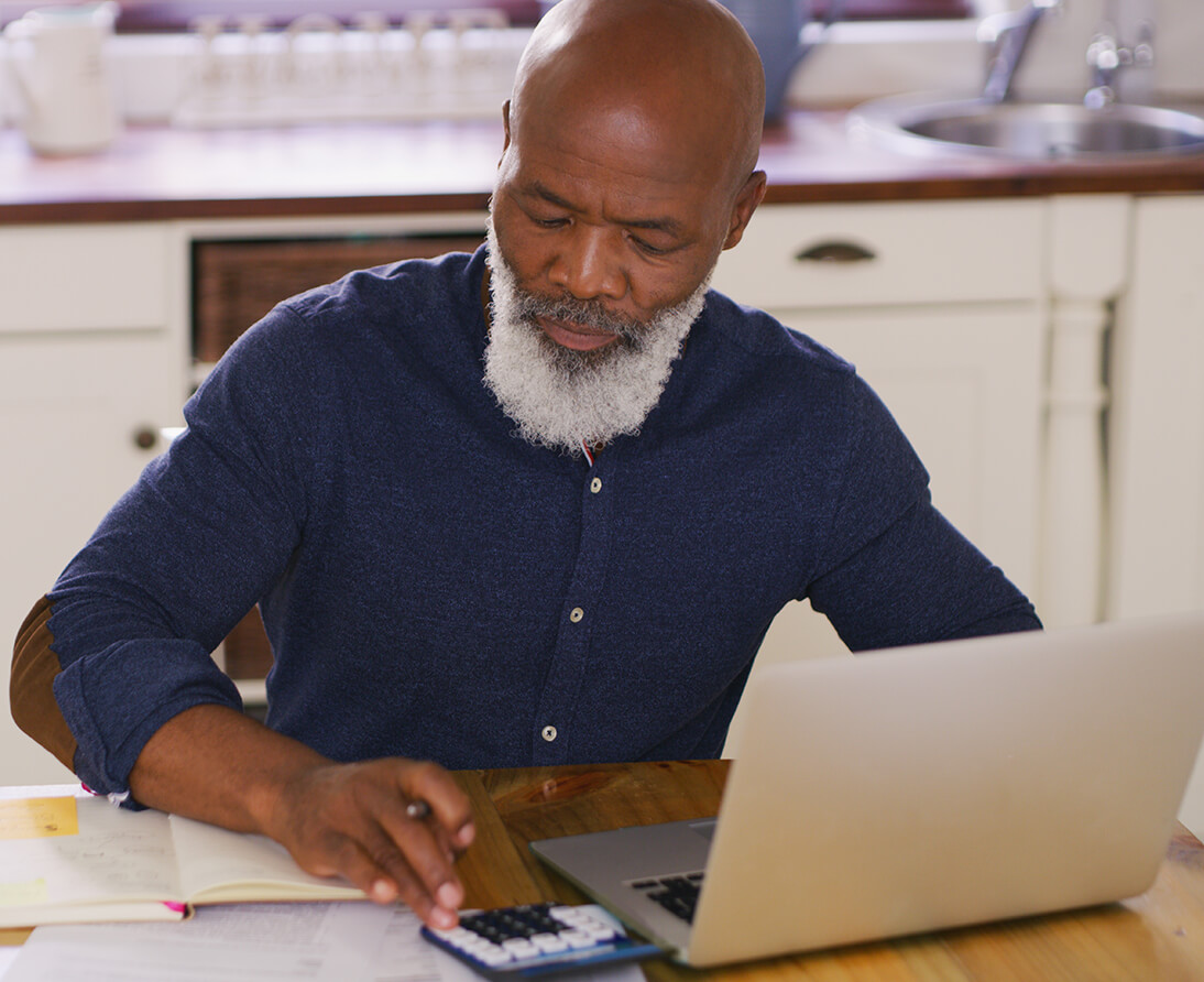 A man sits at a kitchen table with a laptop, typing numbers into a calculator.