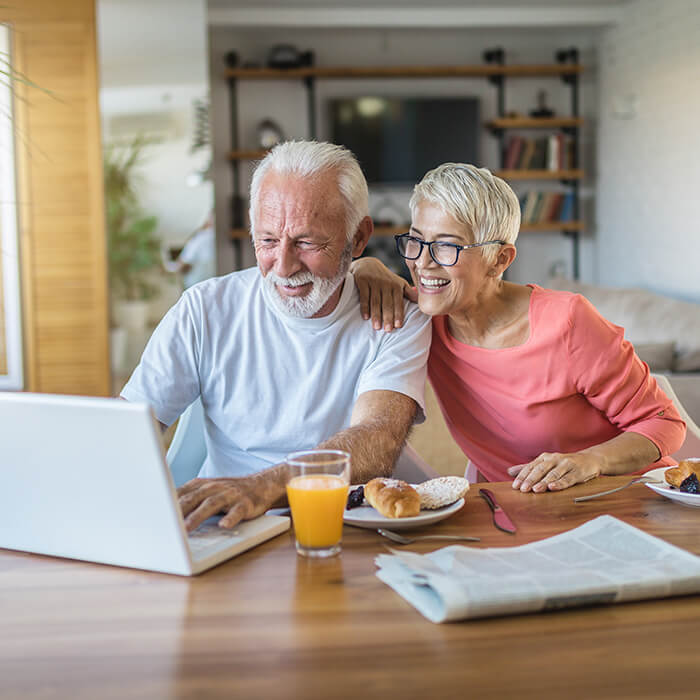 A couple sits at a kitchen table with breakfast food and orange juice, looking at a laptop computer.