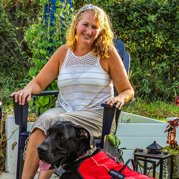 A woman sits on a patio chair, smiling next to a large, brown service dog.